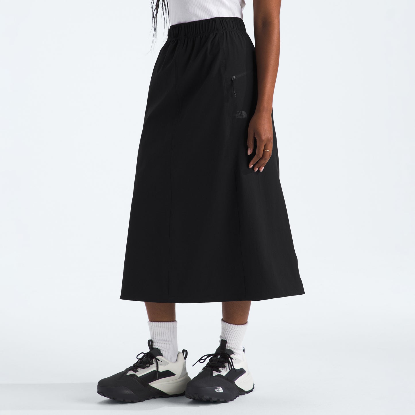 A person in a white top and The North Face W North Dome Wind Skirt with black sneakers and white socks is set against a plain backdrop.