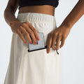 A person wearing the W North Dome Wind Skirt in a light color slips an iPhone into their pocket, while dressed in a black top with visible hands.