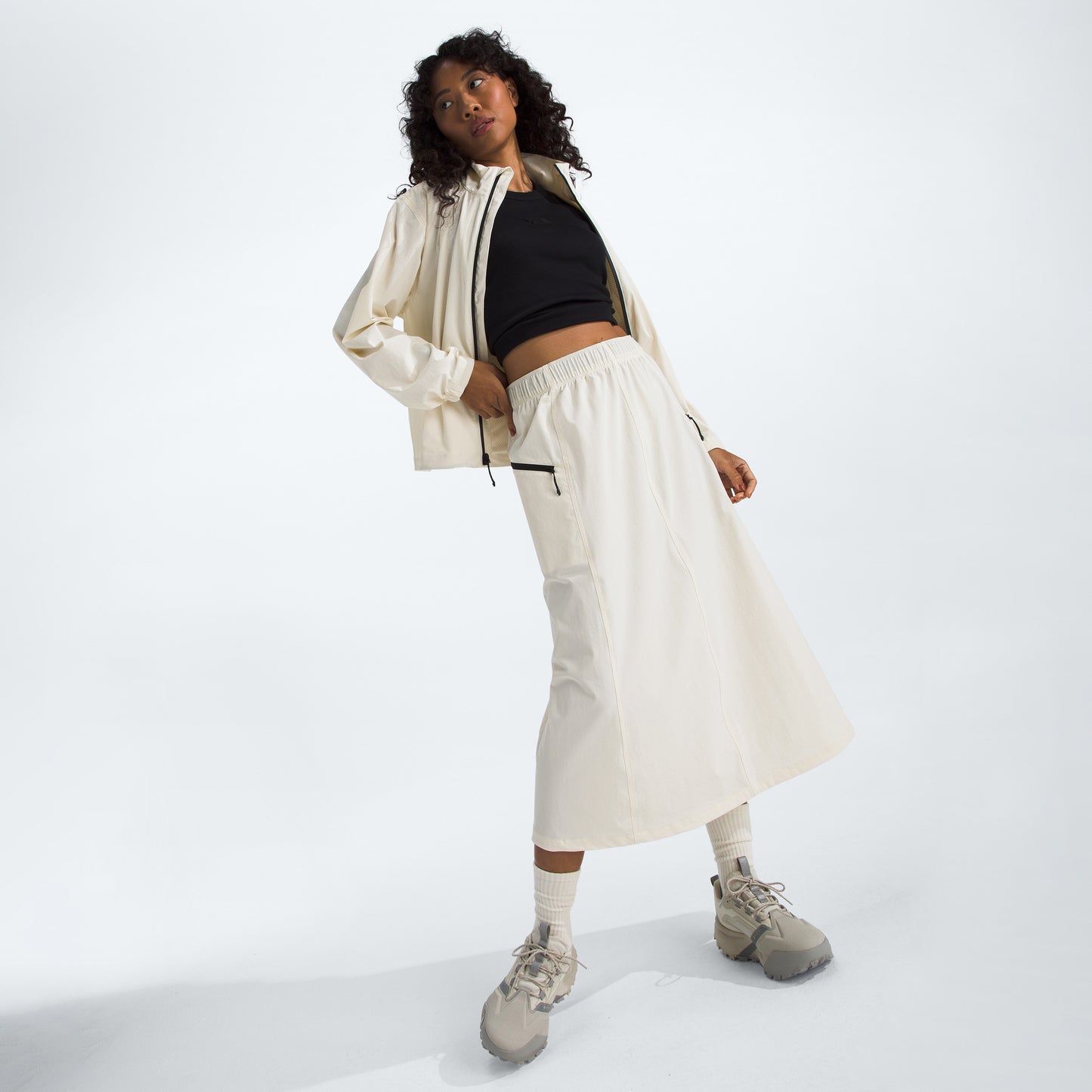 A woman in a cream jacket and The North Face W North Dome Wind Skirt, wearing a black top and beige sneakers, stands with a tilted posture against a plain background.