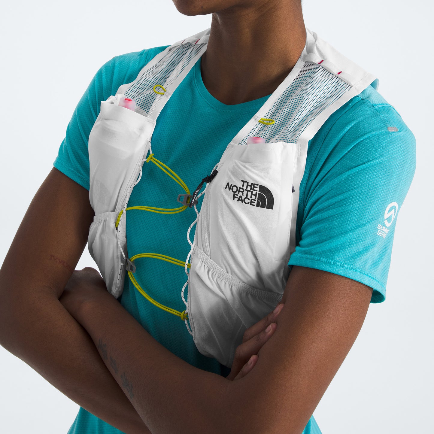 Person wearing a blue short-sleeve shirt and a The North Face U Summit Run Vest 5, featuring neon yellow and pink accents. Arms are confidently crossed, ready for race day.