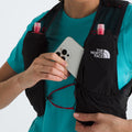 The person in a teal shirt is placing a smartphone into their black U Summit Run Vest 5 by The North Face, with two pink bottles visible in other pockets as they gear up for race day.