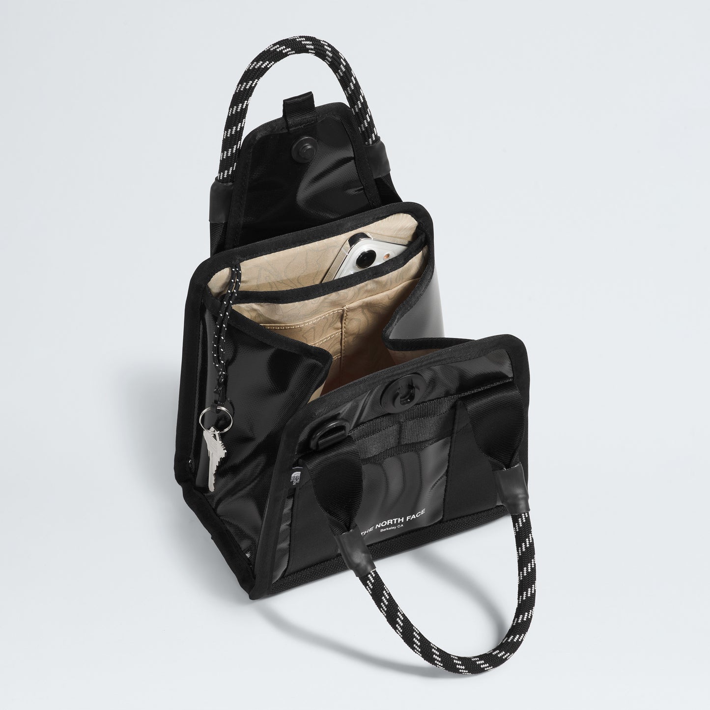 The North Face Base Camp Mini Tote is a black handbag crafted from recycled fabric with white stitching and rope handles. It has a beige-lined interior, smartphone pocket, internal organization with zippered pocket and a key holder, plus an interior strap for modern essentials.