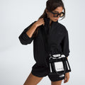 A person in black sunglasses, jacket, and shorts confidently poses against a plain background holding a stylish Base Camp Mini Tote from The North Face, crafted from recycled fabric.