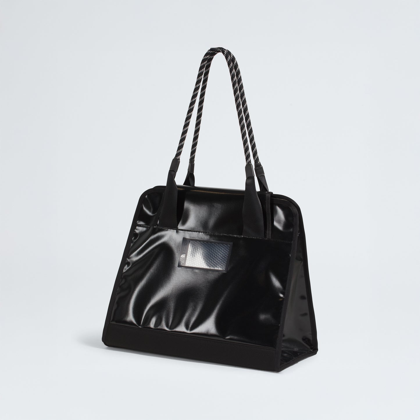 The North Face Base Camp Tote Bag is a durable, black, glossy tote with rope handles and a small rectangular transparent pocket on the front, shown against a plain white background.