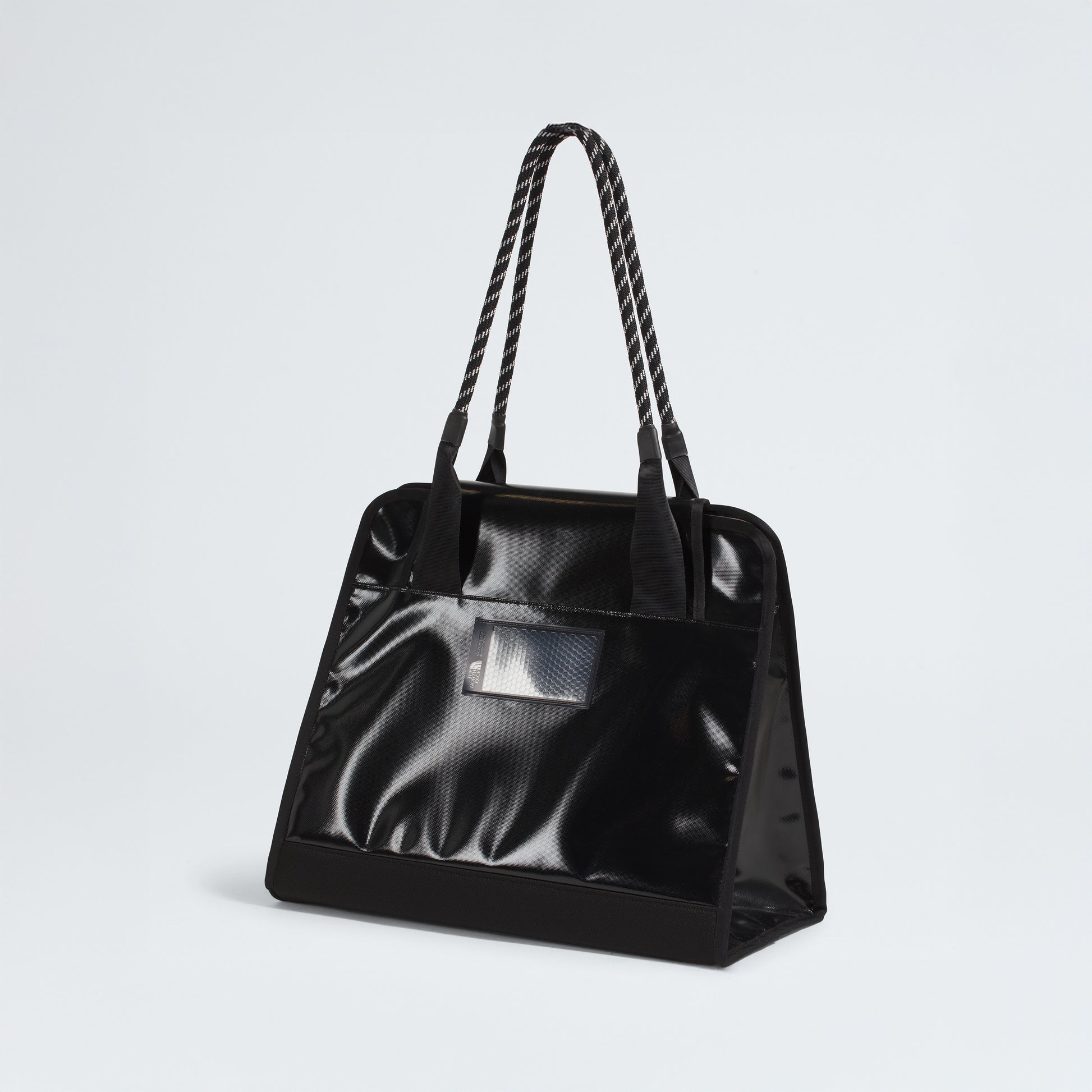 The North Face Base Camp Tote Bag is a durable, black, glossy tote with rope handles and a small rectangular transparent pocket on the front, shown against a plain white background.