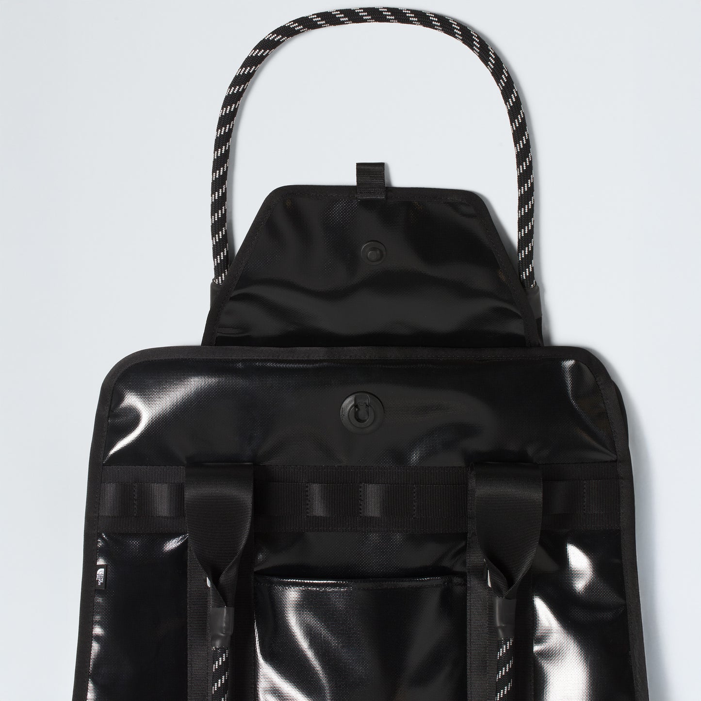 The North Face Base Camp Tote Bag is shown in black with a glossy finish, flap and snap closure, two front straps, braided rope handles, and smart internal organization—offering durable style against a pale background.