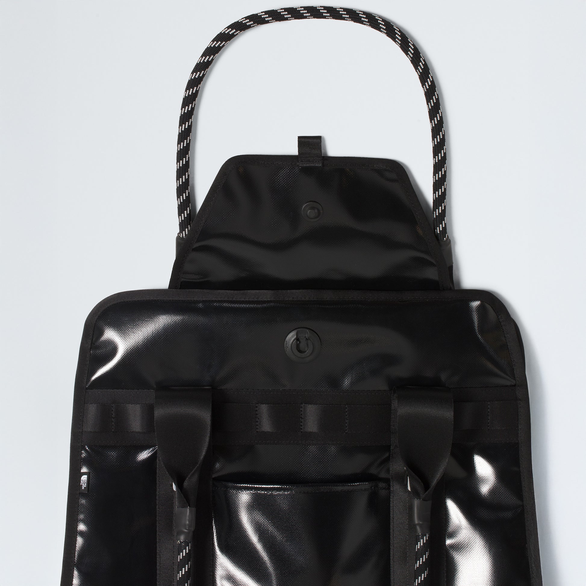 The North Face Base Camp Tote Bag is shown in black with a glossy finish, flap and snap closure, two front straps, braided rope handles, and smart internal organization—offering durable style against a pale background.