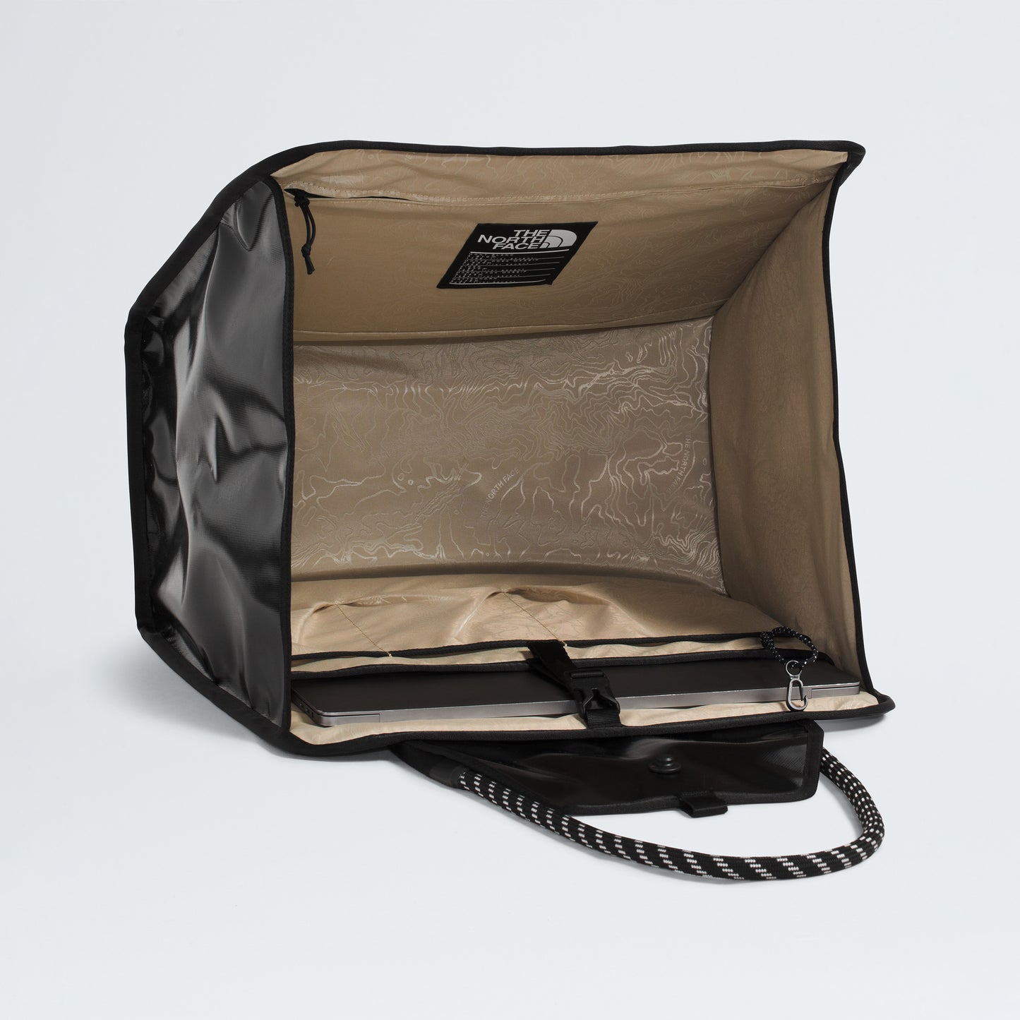 The North Face Base Camp Tote Bag, shown in black and beige with an open front, reveals internal organization pockets and a flat base. A sturdy carry handle and durable construction make it perfect for everyday storage.