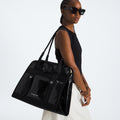 A person in sunglasses, a sleeveless black top, and white pants carries The North Face Base Camp Tote Bag—large and black with smart internal organization—over their shoulder.