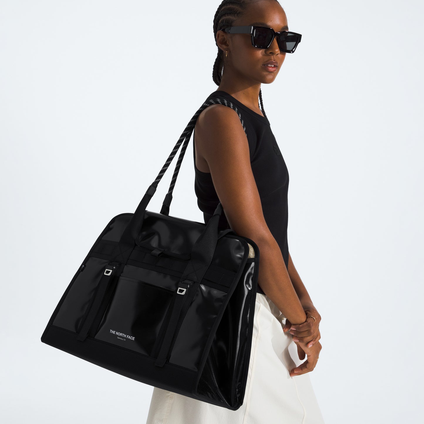 A person in sunglasses, a sleeveless black top, and white pants carries The North Face Base Camp Tote Bag—large and black with smart internal organization—over their shoulder.