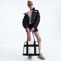 A person stands against a white background wearing sunglasses, a black puffer jacket, white tank top, black shorts, sneakers, and holds The North Face Base Camp Tote Bag, known for its durability and excellent internal organization.