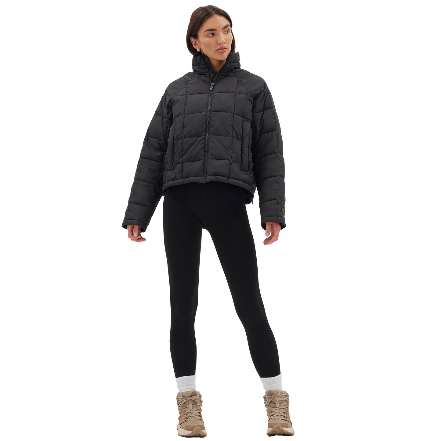 A person wearing a Halfdays Nellie Packable Puffer Jacket, black leggings, and tan boots stands with hands on hips against a white background, ready for outdoor adventure.