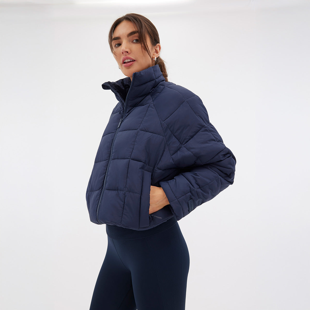 Wearing the Nellie Packable Puffer Jacket by Halfdays and dark leggings, a person stands against a plain white background, prepared for any outdoor adventure.