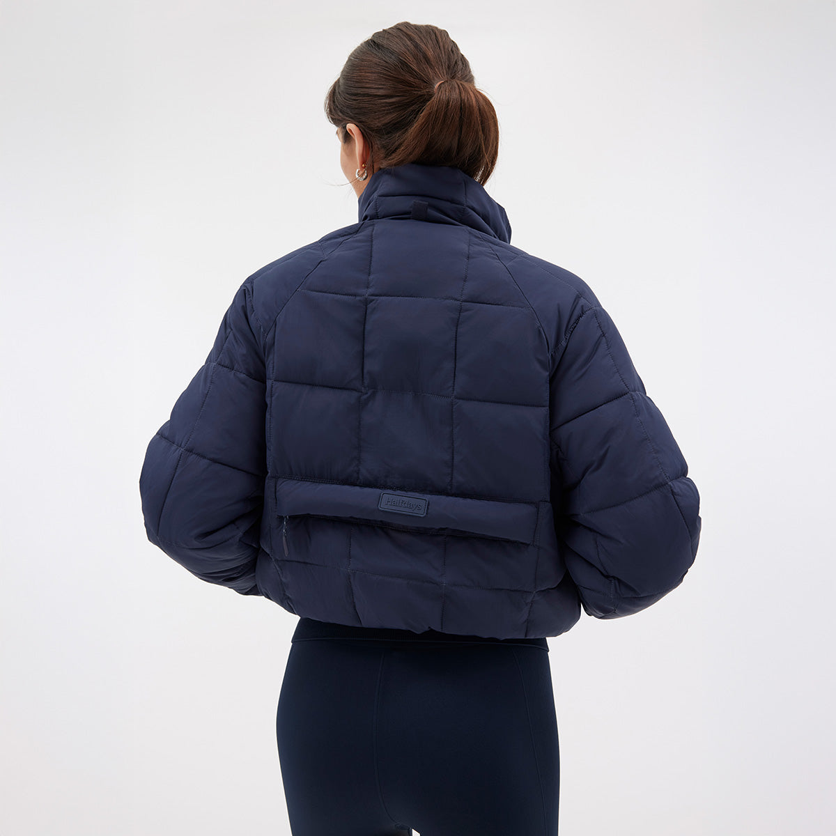 A person in a navy blue Nellie Packable Puffer Jacket by Halfdays, with their back turned, stands against a plain background, ready for outdoor adventure.