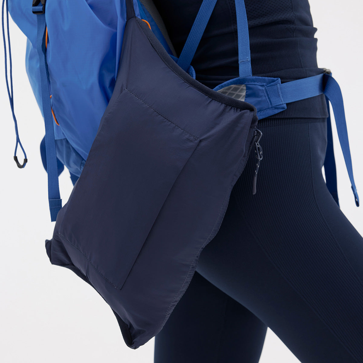 A person ready for an outdoor adventure wears a Nellie Packable Puffer Jacket from Halfdays in dark blue, paired with a blue backpack. The look is completed with a navy pouch on the side strap, perfect for essentials like a travel pillow.