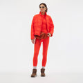 Wearing an orange Nellie Packable Puffer Jacket by Halfdays and matching leggings, along with sunglasses and brown boots, the person stands against a plain white background. A brown waist bag completes their look, making them ready for an outdoor adventure.