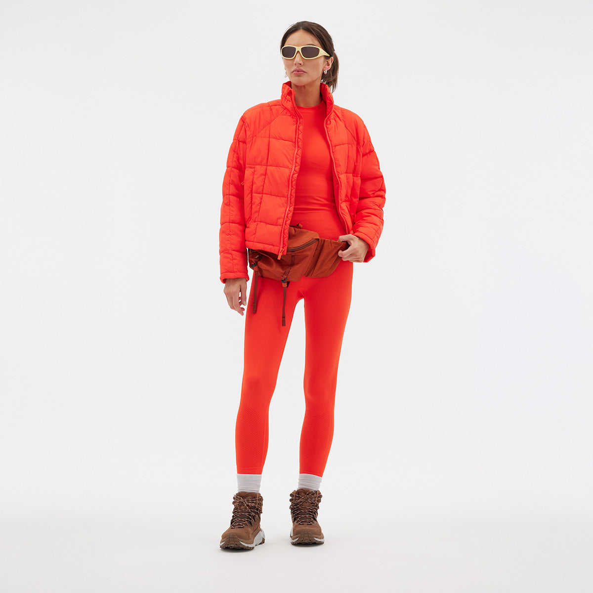 Wearing an orange Nellie Packable Puffer Jacket by Halfdays and matching leggings, along with sunglasses and brown boots, the person stands against a plain white background. A brown waist bag completes their look, making them ready for an outdoor adventure.