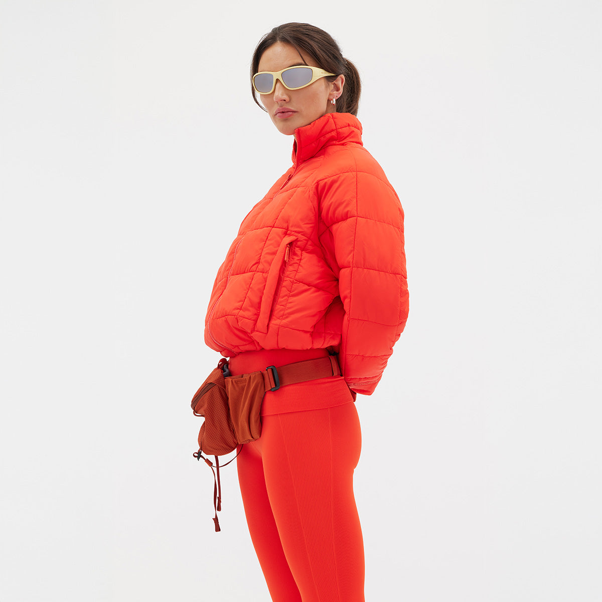 Person in a bright orange Nellie Packable Puffer Jacket by Halfdays, matching leggings, sunglasses, and a small bag on a white background.