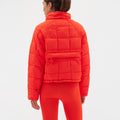 Clad in a vibrant Nellie Packable Puffer Jacket by Halfdays, an individual stands with matching leggings, facing away and ready for adventure, a small backpack poised on the jacket.