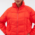 An individual dons the Nellie Packable Puffer Jacket by Halfdays, featuring a bright orange color, lightweight quilting, high collar, and zippered closure, while standing against a plain background.
