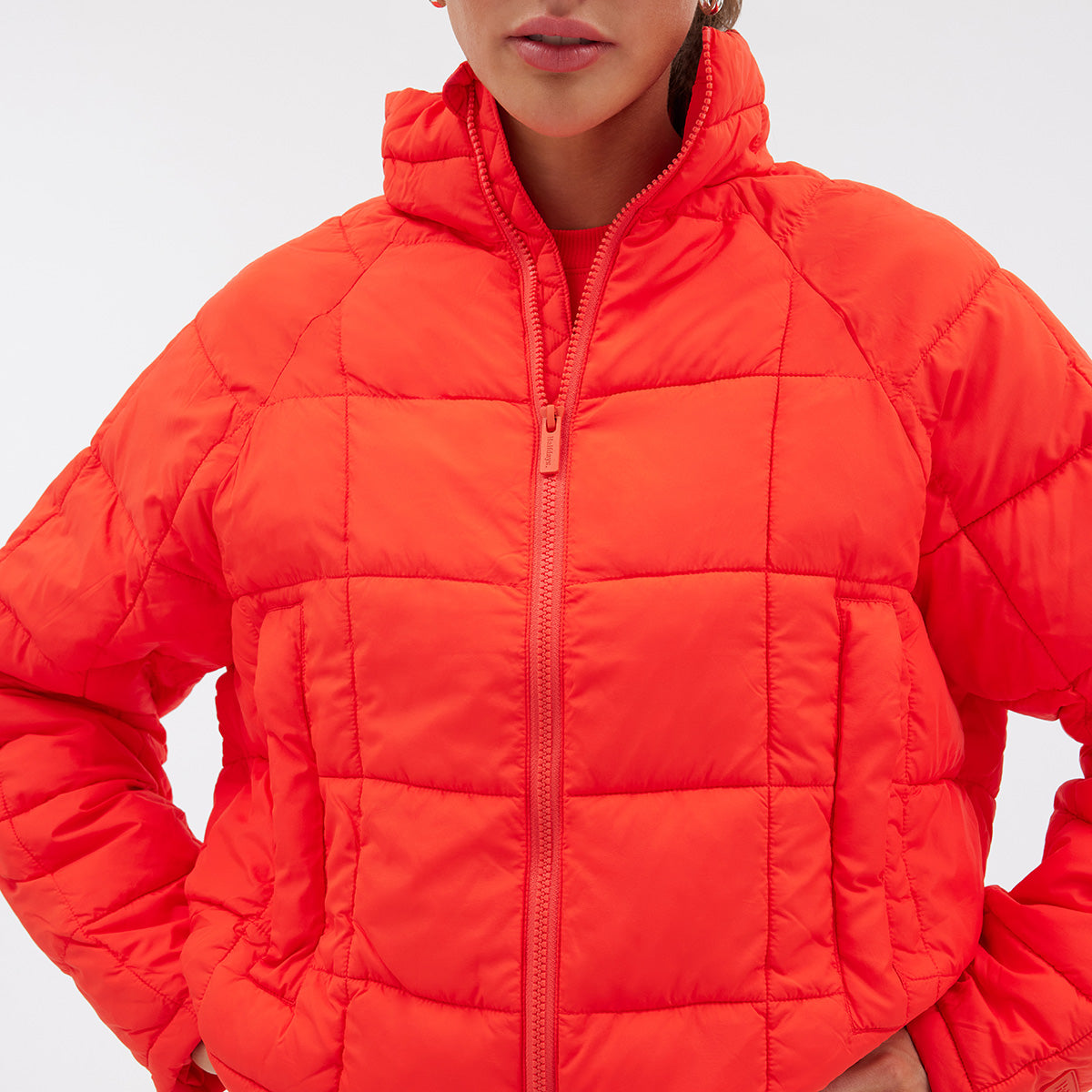 An individual dons the Nellie Packable Puffer Jacket by Halfdays, featuring a bright orange color, lightweight quilting, high collar, and zippered closure, while standing against a plain background.