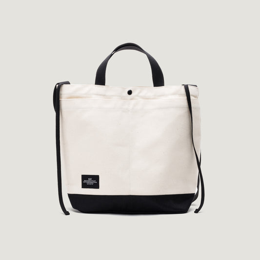 The New York Tote Medium by Bags in Progress is a natural/black canvas bag with a black base and trim. It features two handles, an adjustable shoulder strap, a small black front label, and exterior pockets for easy access to essentials.