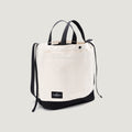The New York Tote Medium by Bags in Progress is a natural/black beige canvas tote with black handles, trim, a front pocket, and a black label with white text. It also includes exterior pockets for added convenience.