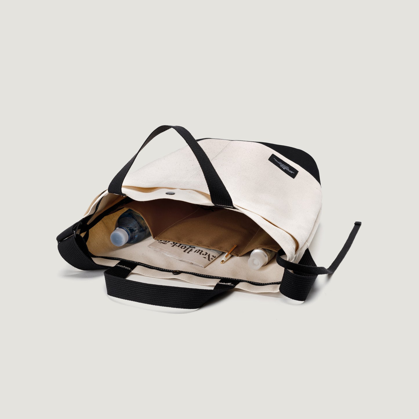 Open New York Tote Medium, Natural/Black from Bags in Progress on a light background, displaying contents like a newspaper, a white bottle, and a clear plastic bottle inside.