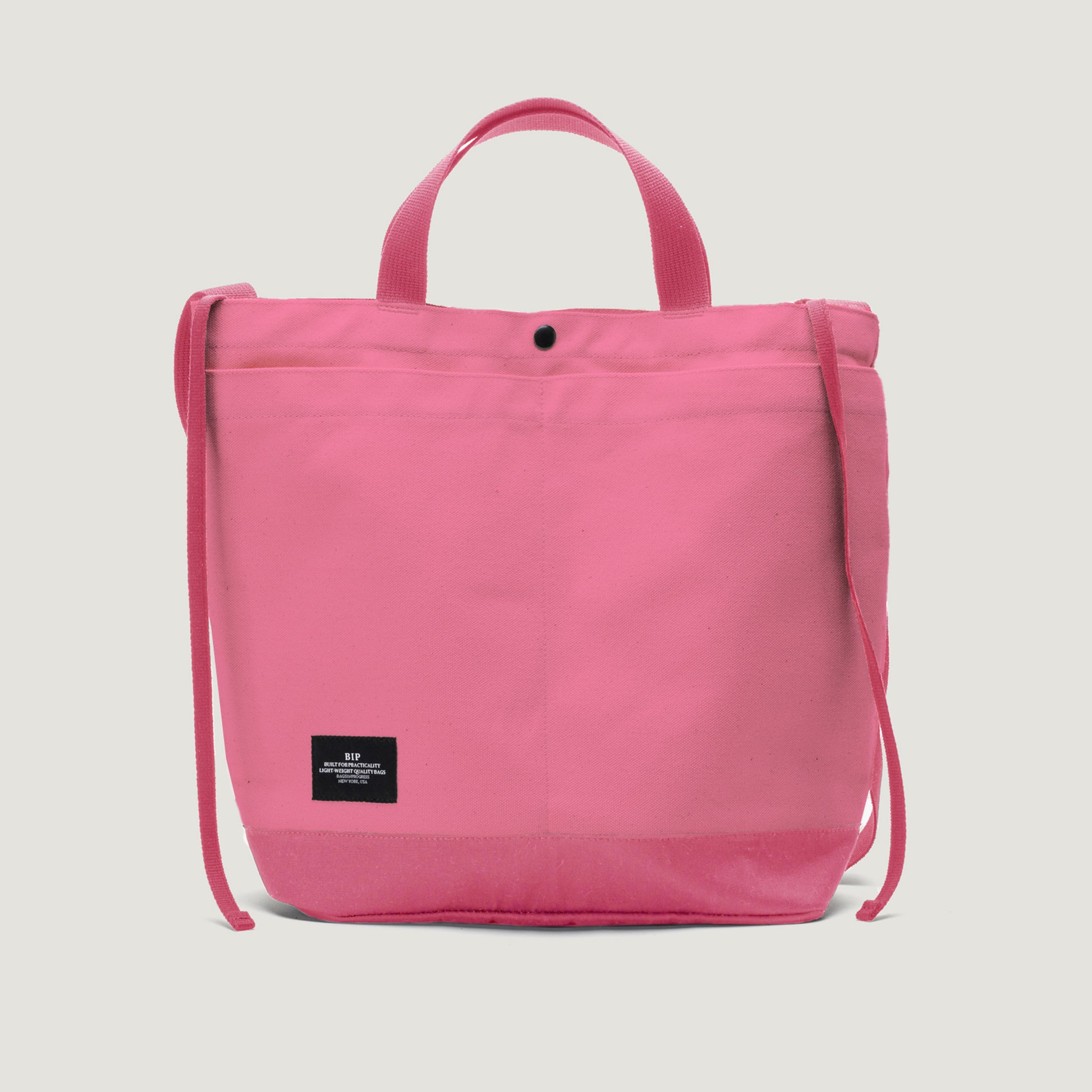 The New York Tote Medium, Special Edition: Candy Pink by Bags in Progress is a pink tote with short handles, an adjustable shoulder strap, a front pocket, open top, and a small black label near the bottom.