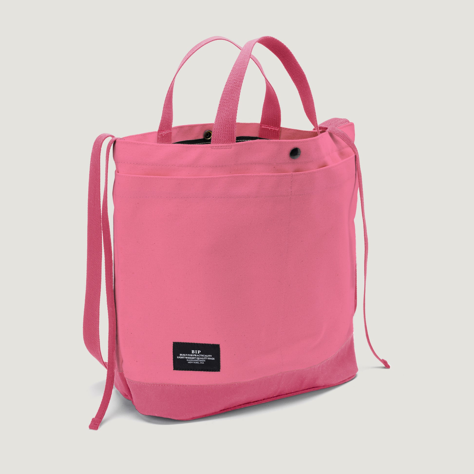 The Bags in Progress New York Tote Medium, Special Edition: Candy Pink, features an open top, two short handles, adjustable shoulder straps, and a black front label, shown against a white background.