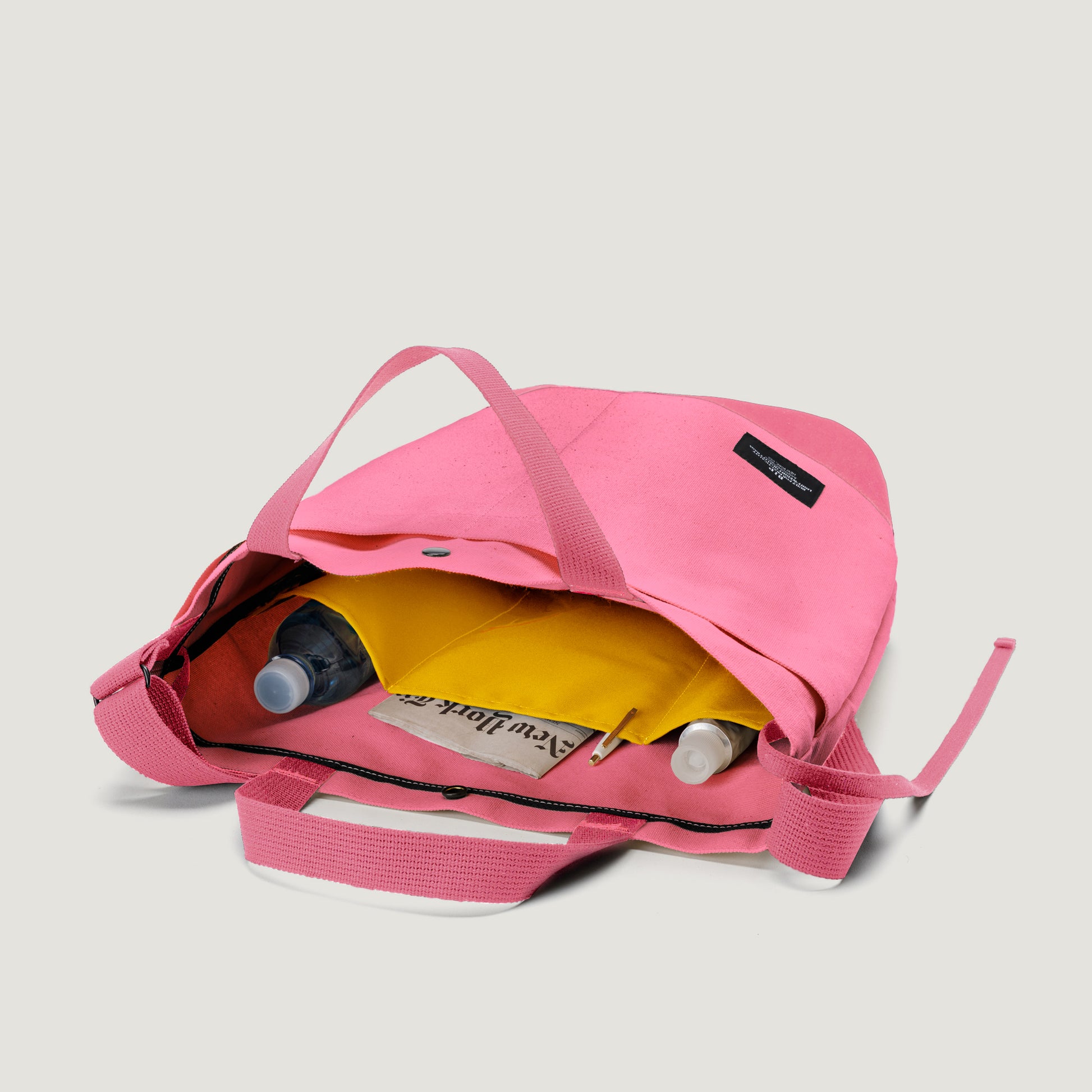 The Bags in Progress New York Tote Medium, Special Edition: Candy Pink, features an open top, yellow interior pocket, and is shown with a water bottle, tube, and wallet on a plain background.