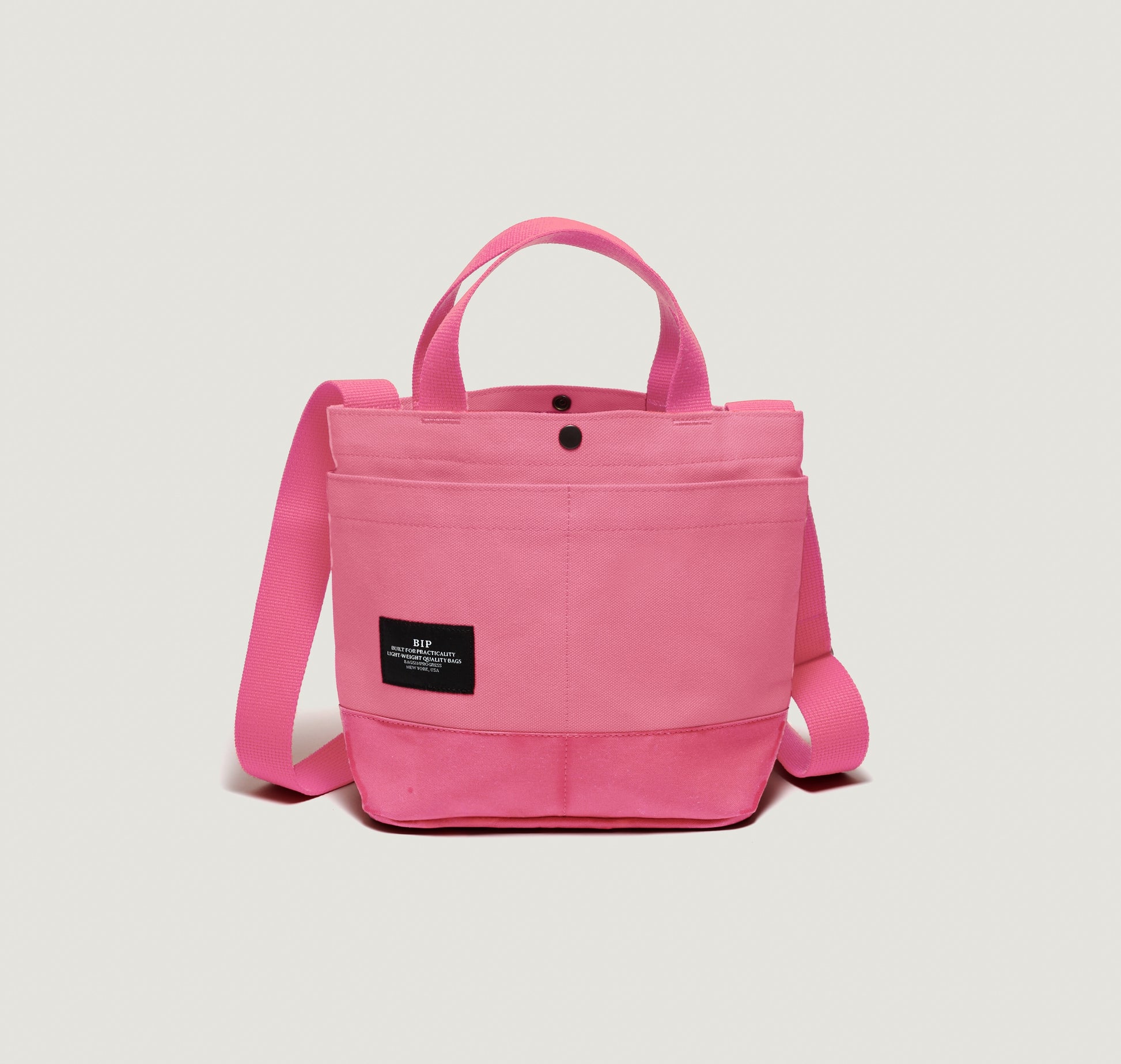 The Bags in Progress New York Tote Small, Special Edition: Candy Pink, features dual handles, exterior pockets, an adjustable shoulder strap, and a black label on the front pocket, shown on a plain white background.