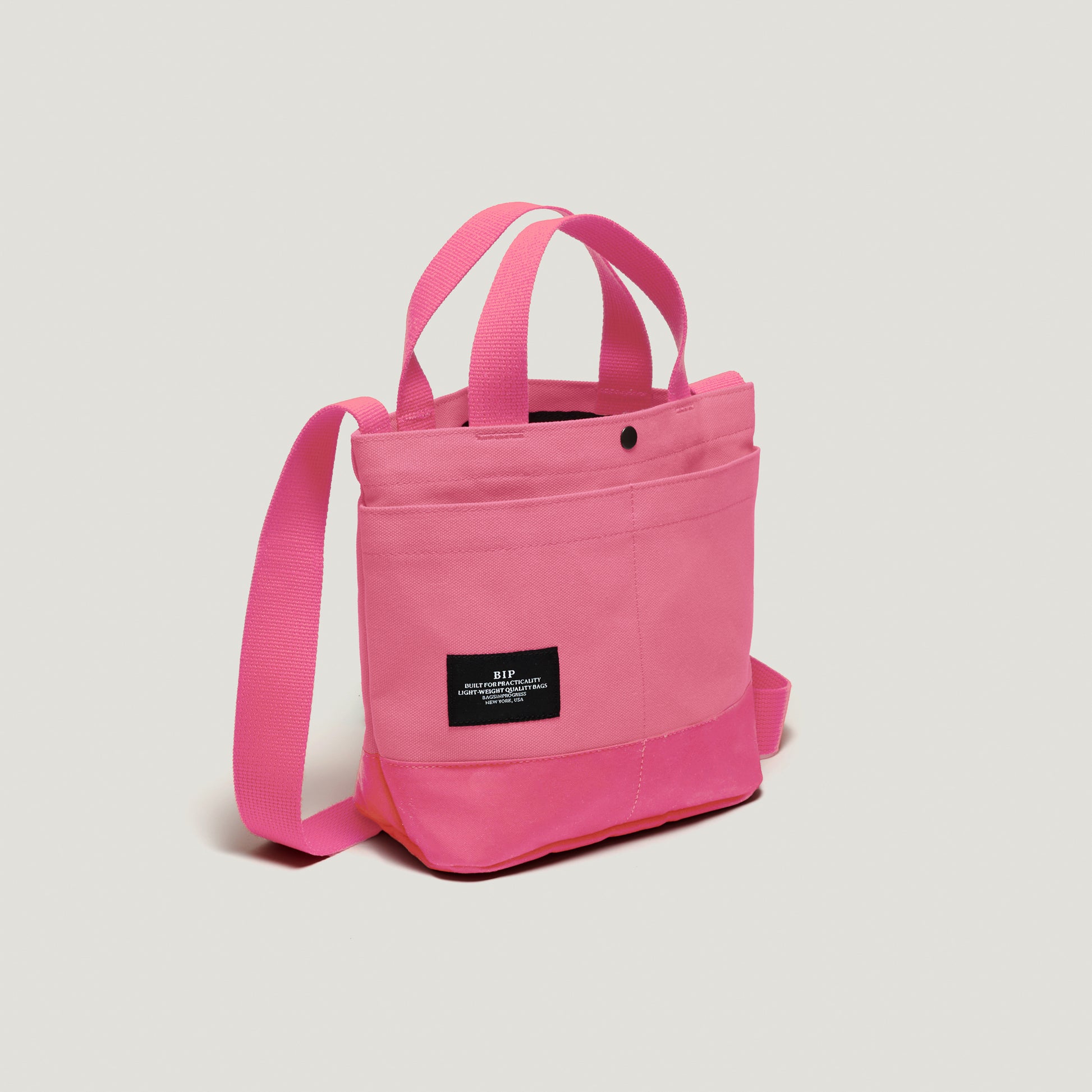 The New York Tote Small, Special Edition: Candy Pink by Bags in Progress is a pink tote with dual handles, exterior pockets, an adjustable shoulder strap, and a sleek black rectangular label on the front.