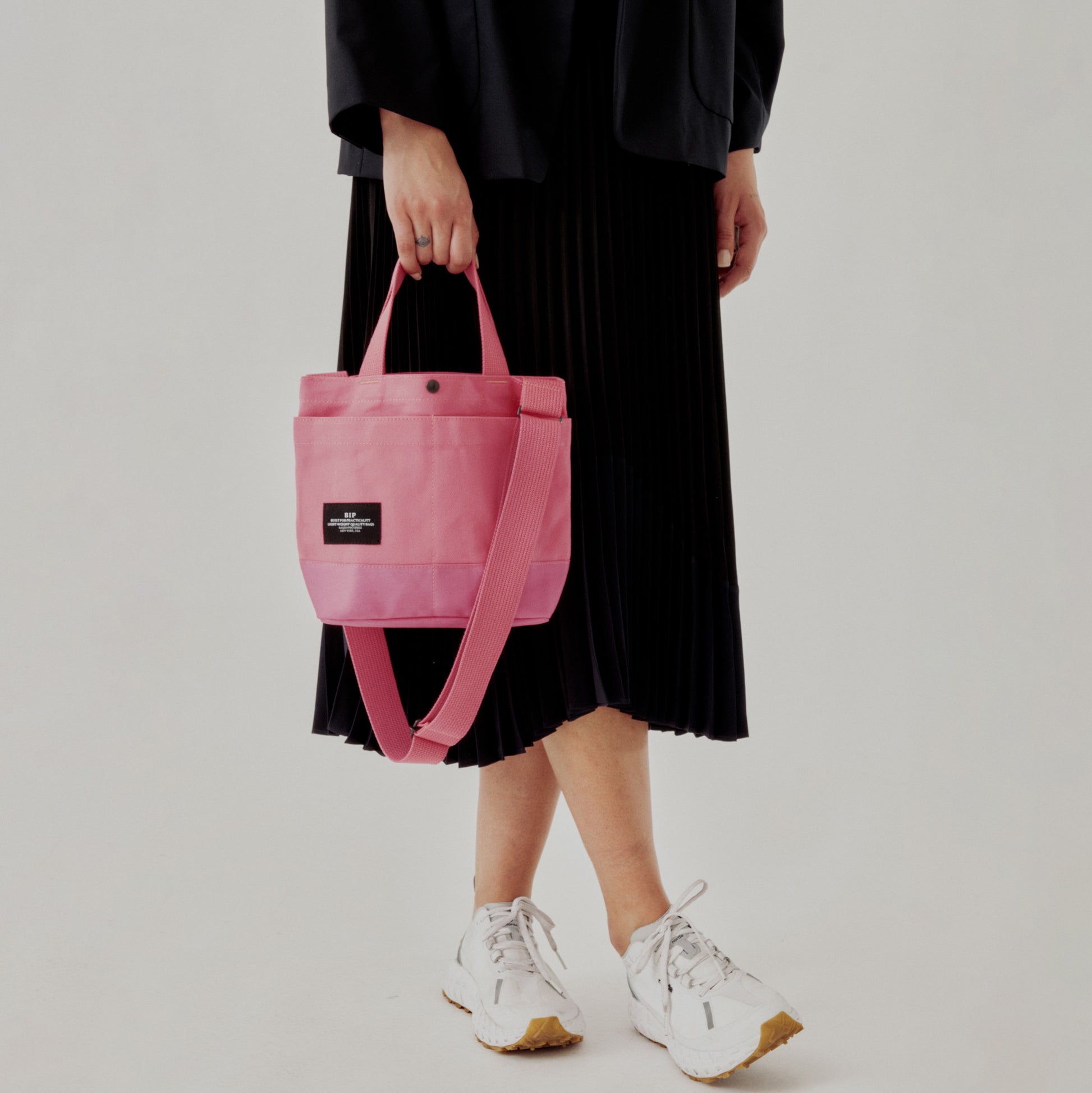 A person in a black skirt and white sneakers holds the Bags in Progress New York Tote Small, Special Edition: Candy Pink, featuring a black label and exterior pockets, against a plain background.