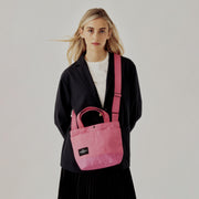 A woman in a black blazer and white shirt stands against a plain background, carrying the Bags in Progress New York Tote Small, Special Edition: Candy Pink, featuring handles, exterior pockets, and a black label.
