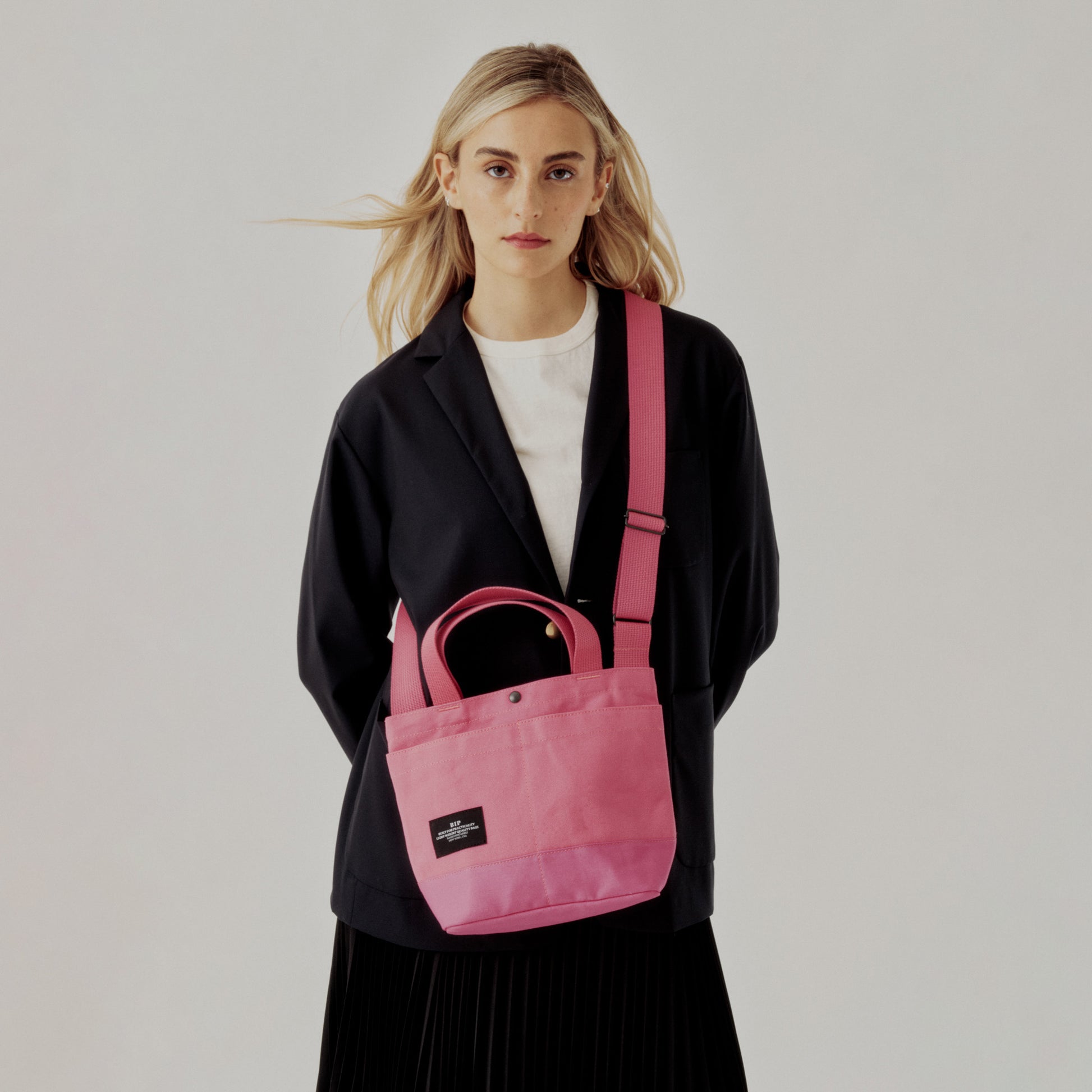 A woman in a black blazer and white shirt stands against a plain background, carrying the Bags in Progress New York Tote Small, Special Edition: Candy Pink, featuring handles, exterior pockets, and a black label.