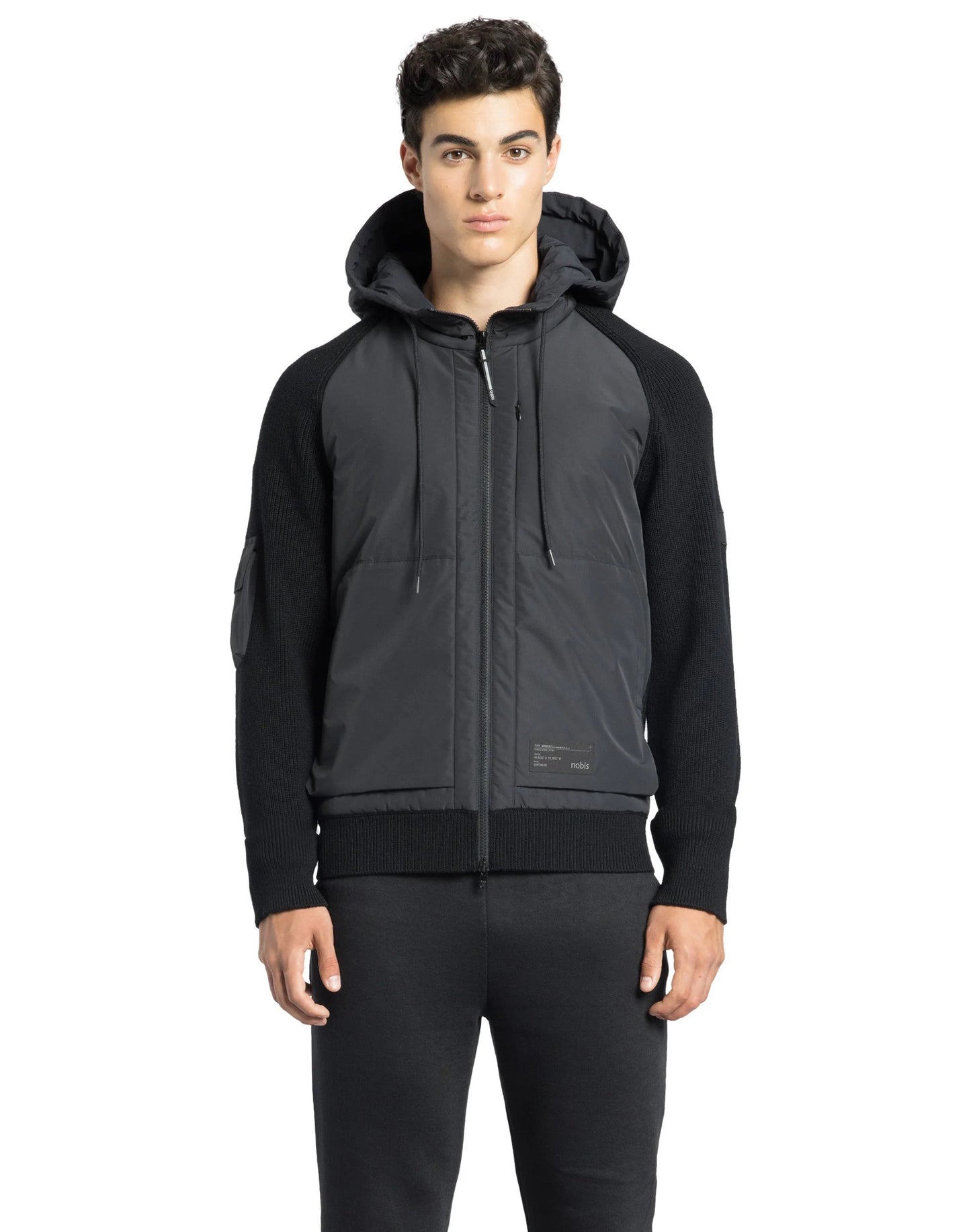 A person wearing a luxurious Hedge Men's Performance Hoodie and dark pants stands against a white background. The hoodie, crafted by Nobis from premium merino wool, effortlessly combines style with water resistance, offering both elegance and practicality.