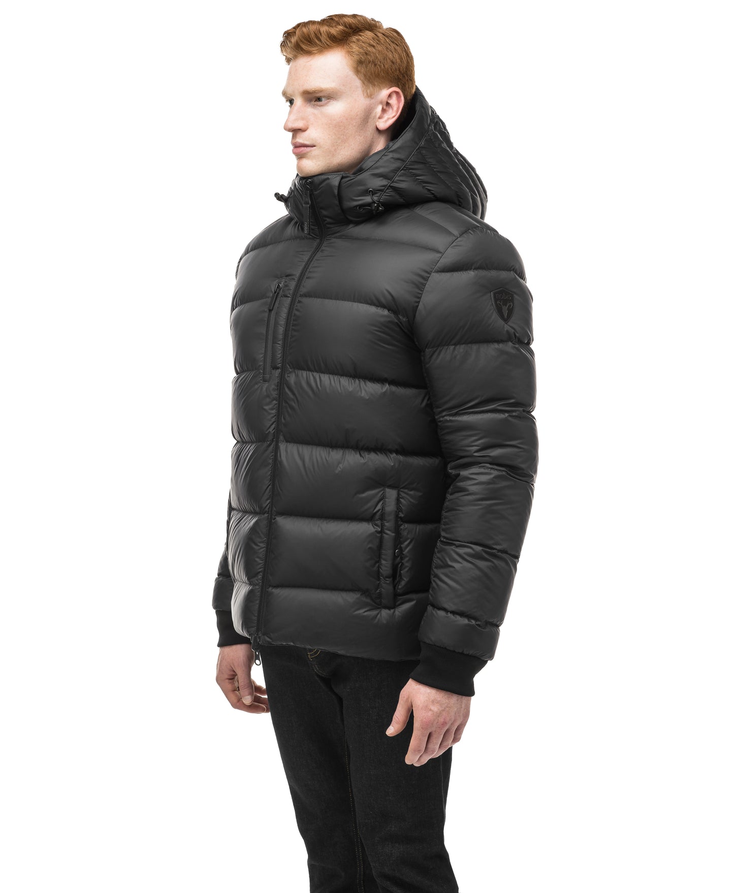 A person wearing the Nobis Oliver Men's Reversible Puffer Jacket in black with a hood stands sideways against a plain white background.