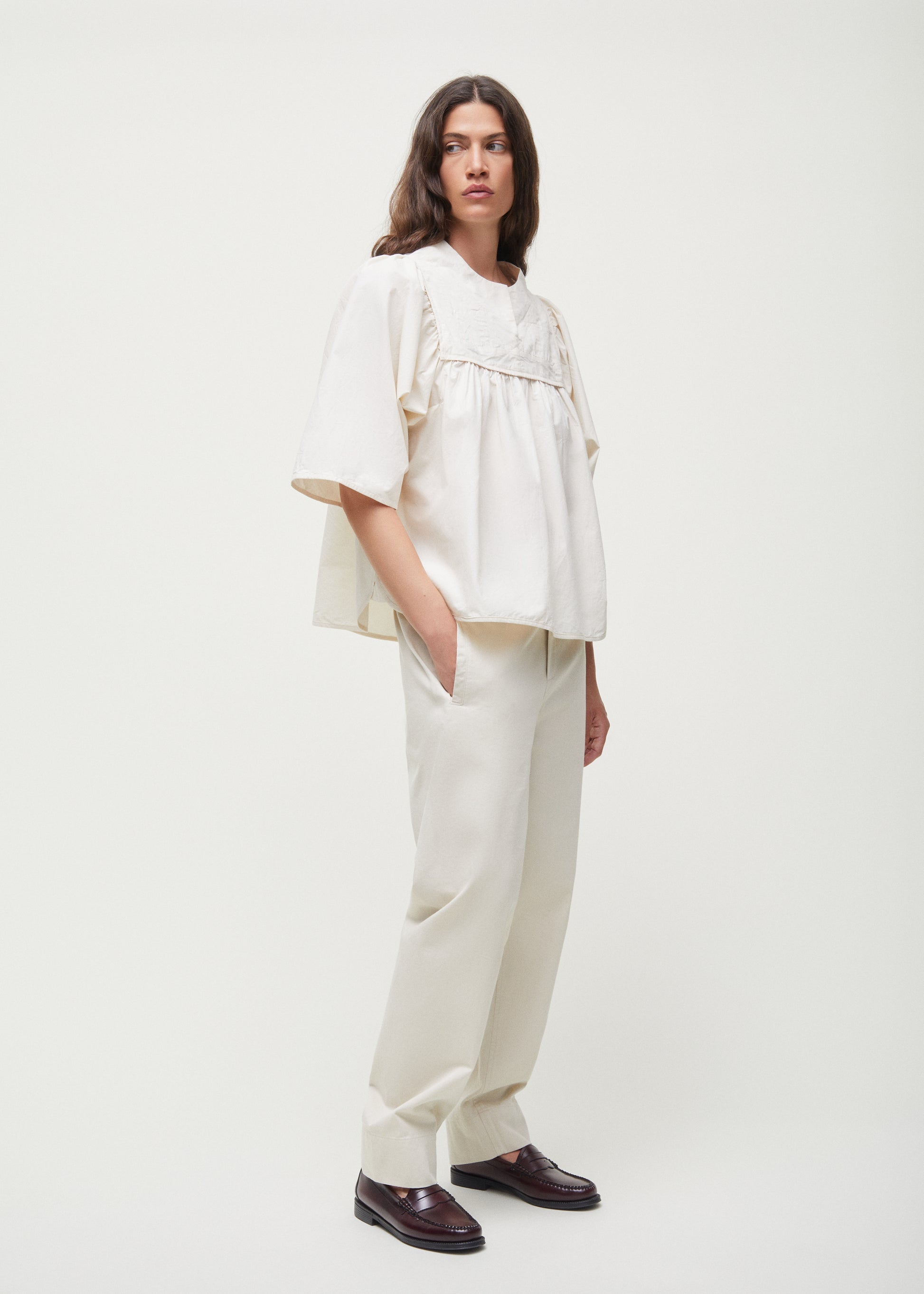 A person with long hair, wearing the loose-fitting Nomi Blouse in beige, crafted by Aiayu from regenerative cotton, paired with matching trousers, stands against a plain background with hands in pockets.