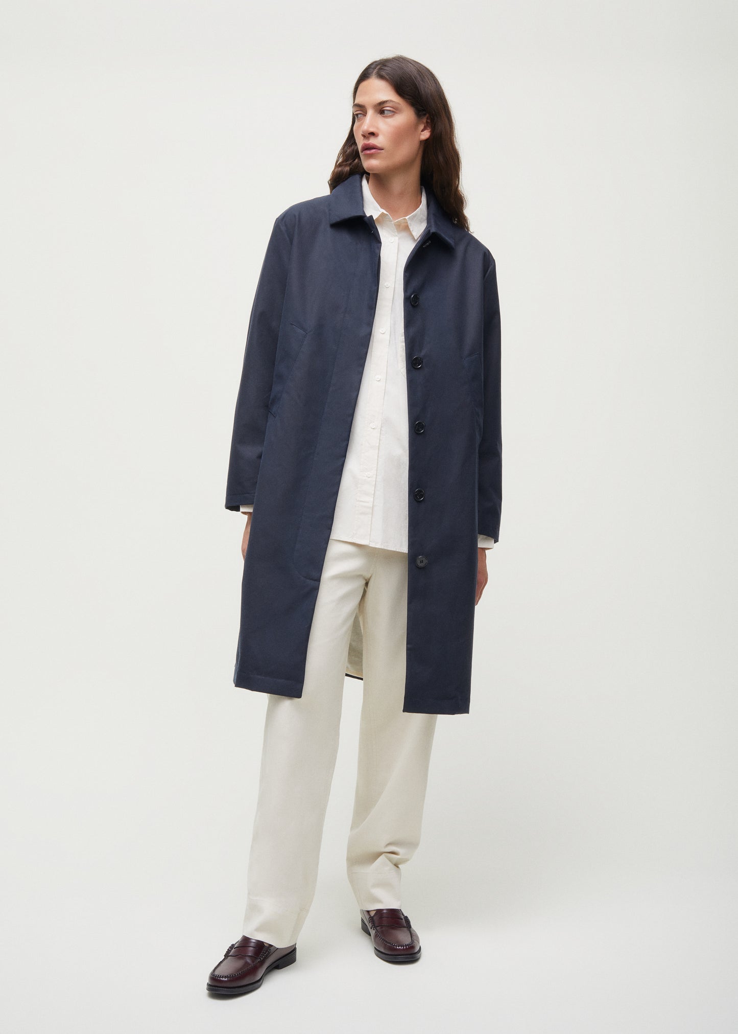 Person wearing Aiayu's Noor Coat Oil in classic navy over a white shirt and cream pants, standing against a plain background; the coat is crafted from wax-coated organic cotton.