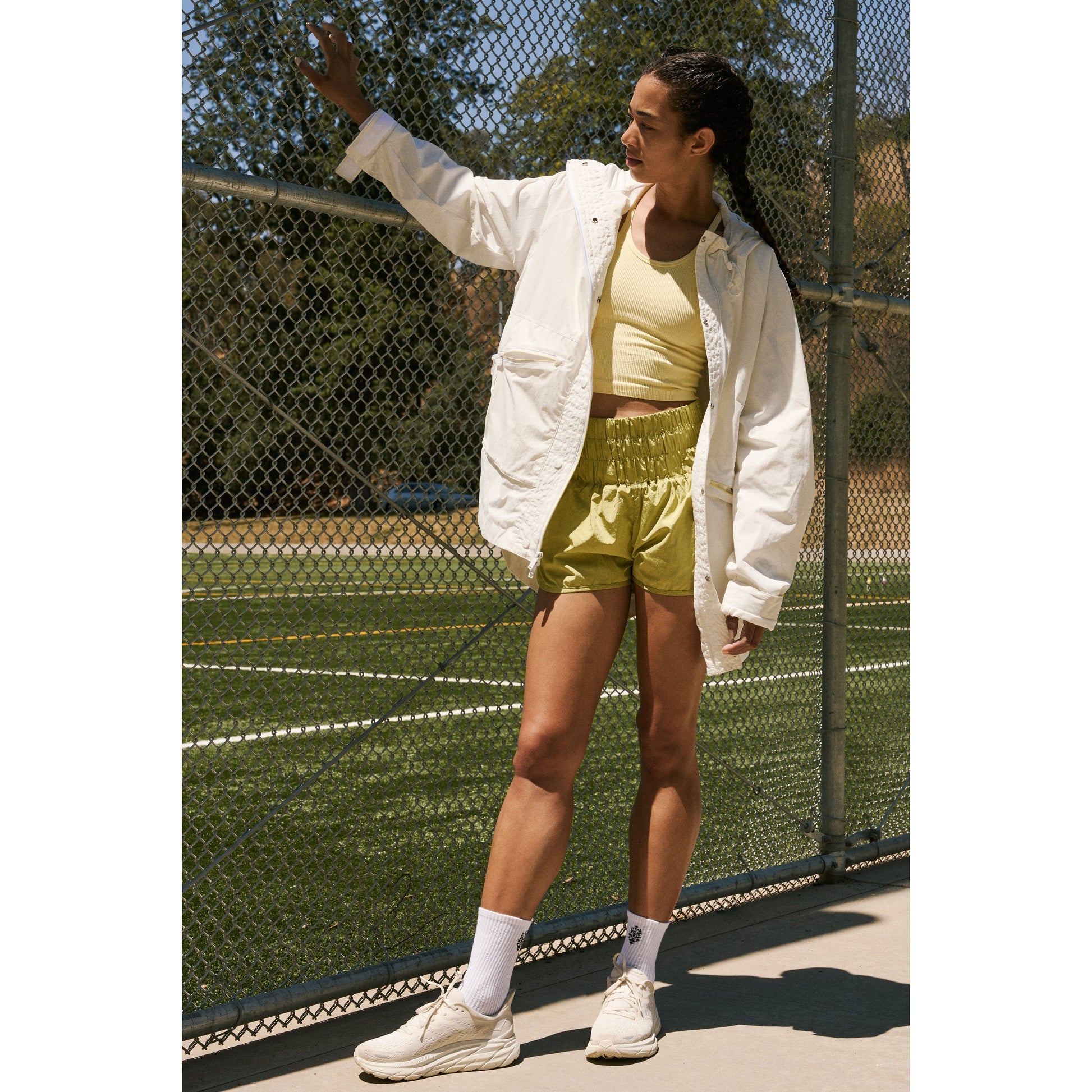 A young woman in sporty attire stands on a tennis court, raising her hand to shield her eyes from the sun. She wears a Singin in the Rain Jacket, Painted White by Free People Movement, ideal for unpredictable weather.