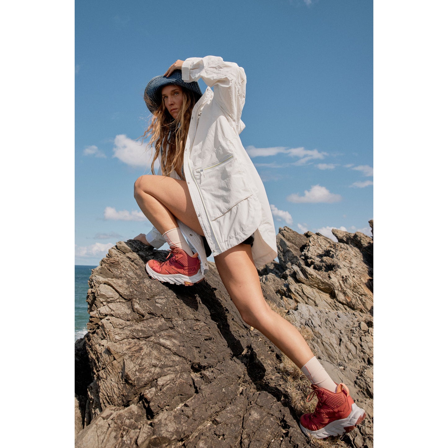 Woman in a Singin in the Rain Jacket, Painted White and red shoes posing on a rocky seaside cliff under a clear blue sky.