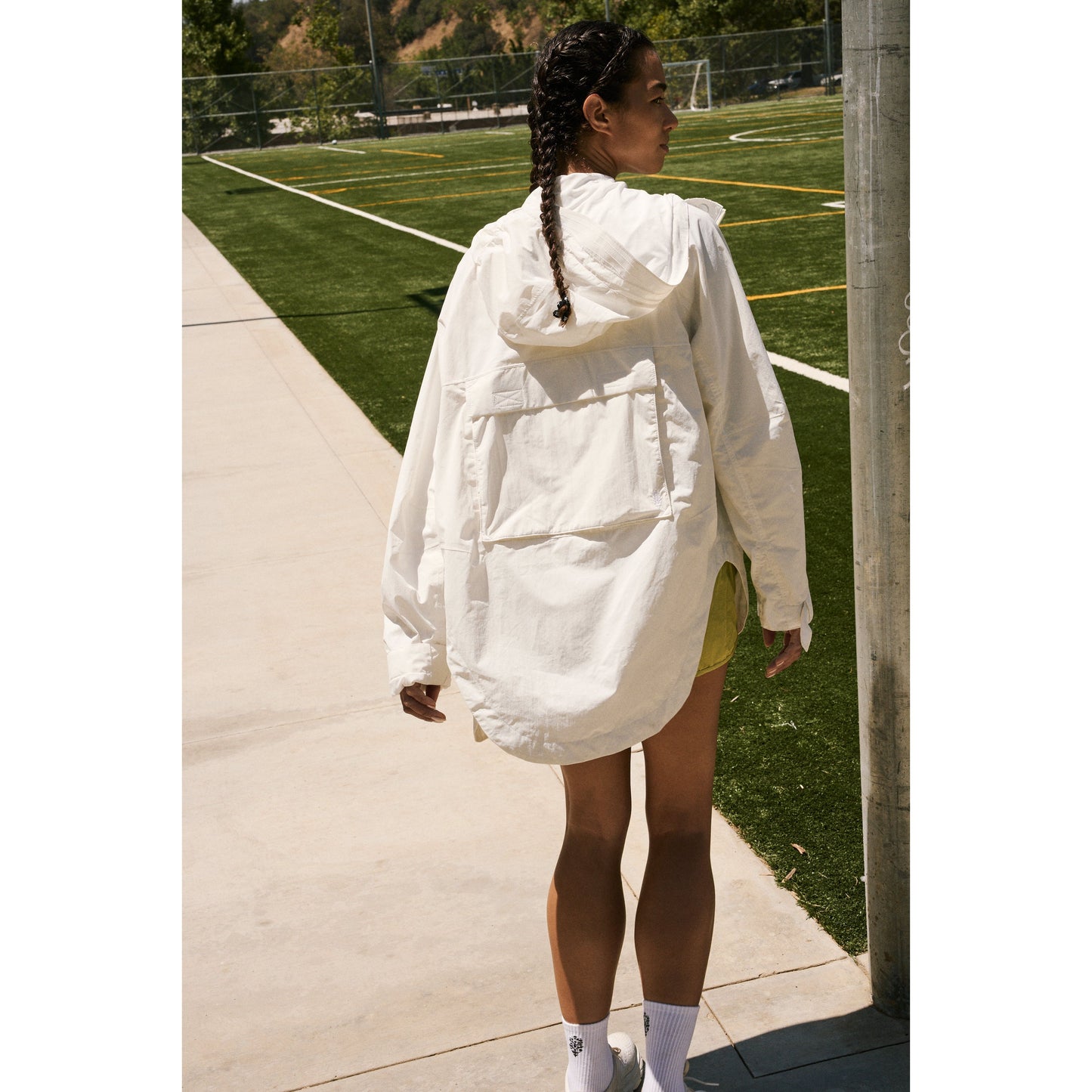 A woman in a Singin in the Rain Jacket by Free People Movement and yellow shorts standing by a pole on a sunny sports field, looking away from the camera.