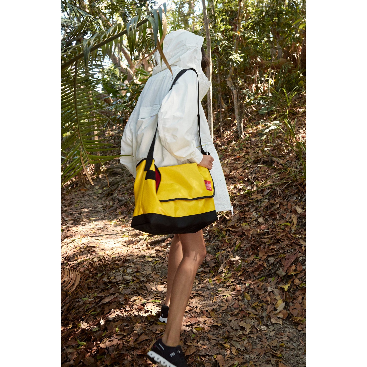 A person in a Singin in the Rain Jacket, Painted White hooded rain jacket and shorts carrying a bright yellow bag walks on a forest path.