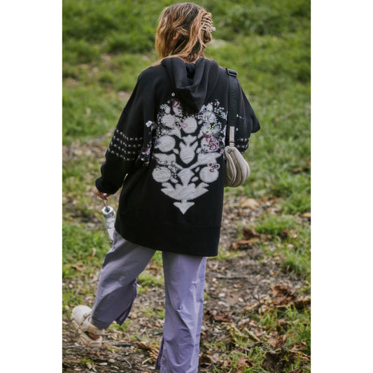 A person walking on grass, viewed from behind, wearing a Free People Movement At My Best Embroidered Sweatshirt in Black Combo with a floral skull design and light purple pants.
