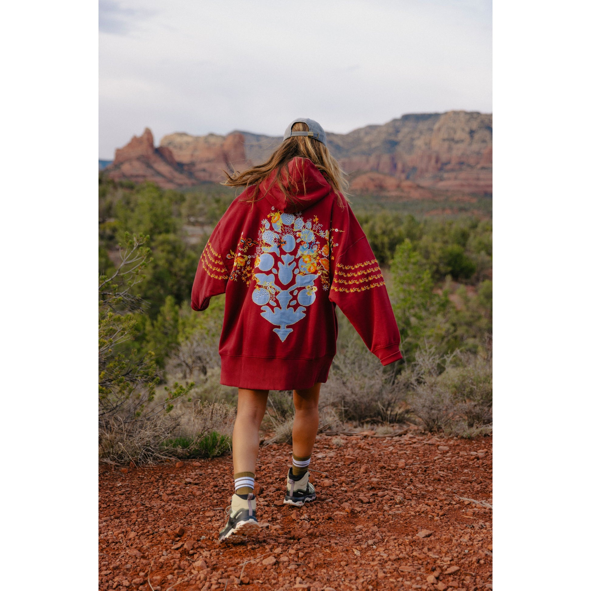 A woman in a Free People Movement At My Best Embroidered Sweatshirt in Sour Cherry Combo and hiking boots walks on a red dirt trail with lush greenery and rocky terrain in the background.