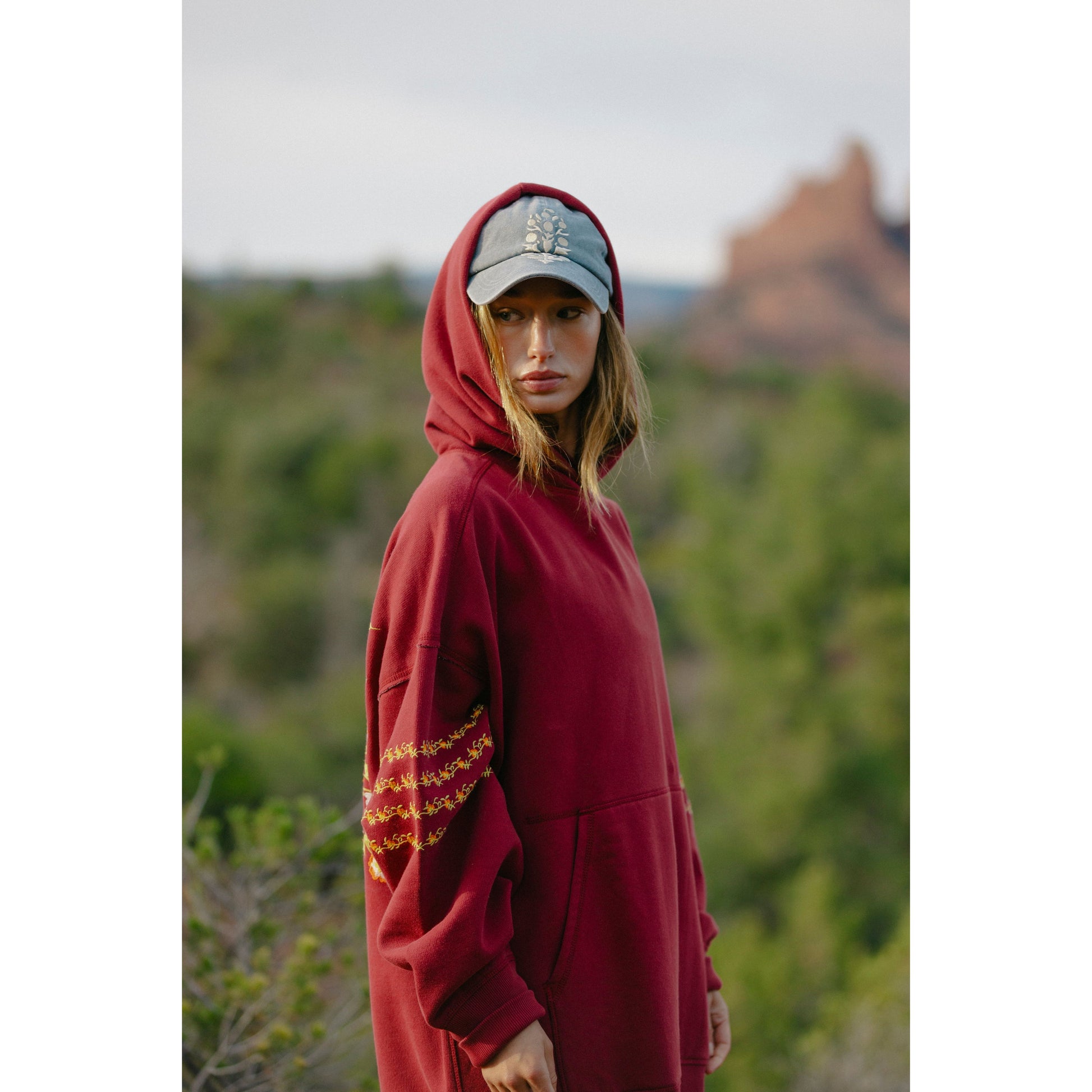 Woman in a red At My Best Embroidered Sweatshirt by Free People Movement with a kangaroo pocket, wearing a baseball cap, stands outdoors with a blurred desert landscape in the background.