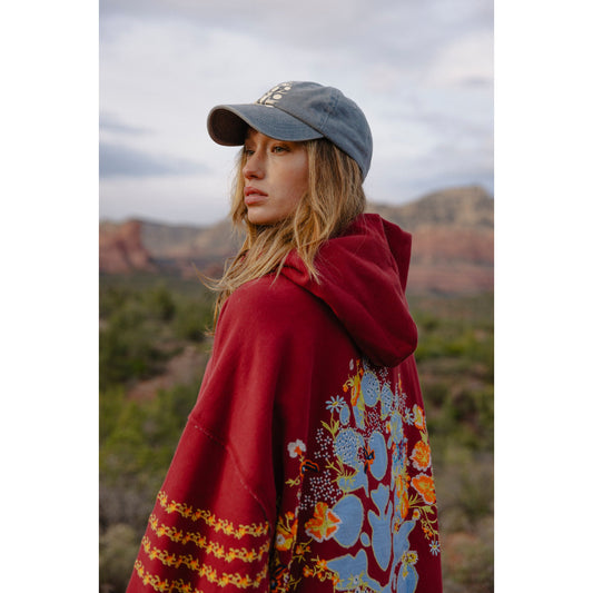 Woman in a Free People Movement At My Best Embroidered Sweatshirt in Sour Cherry Combo and blue cap looking over her shoulder, with a backdrop of desert mountains.