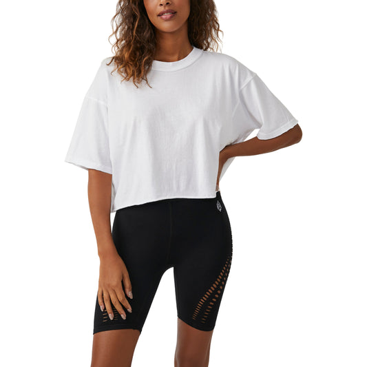 Woman in a white Free People Movement Inspire Tee and black cycling shorts with mesh detail, standing with hands on hips, looking at the camera.