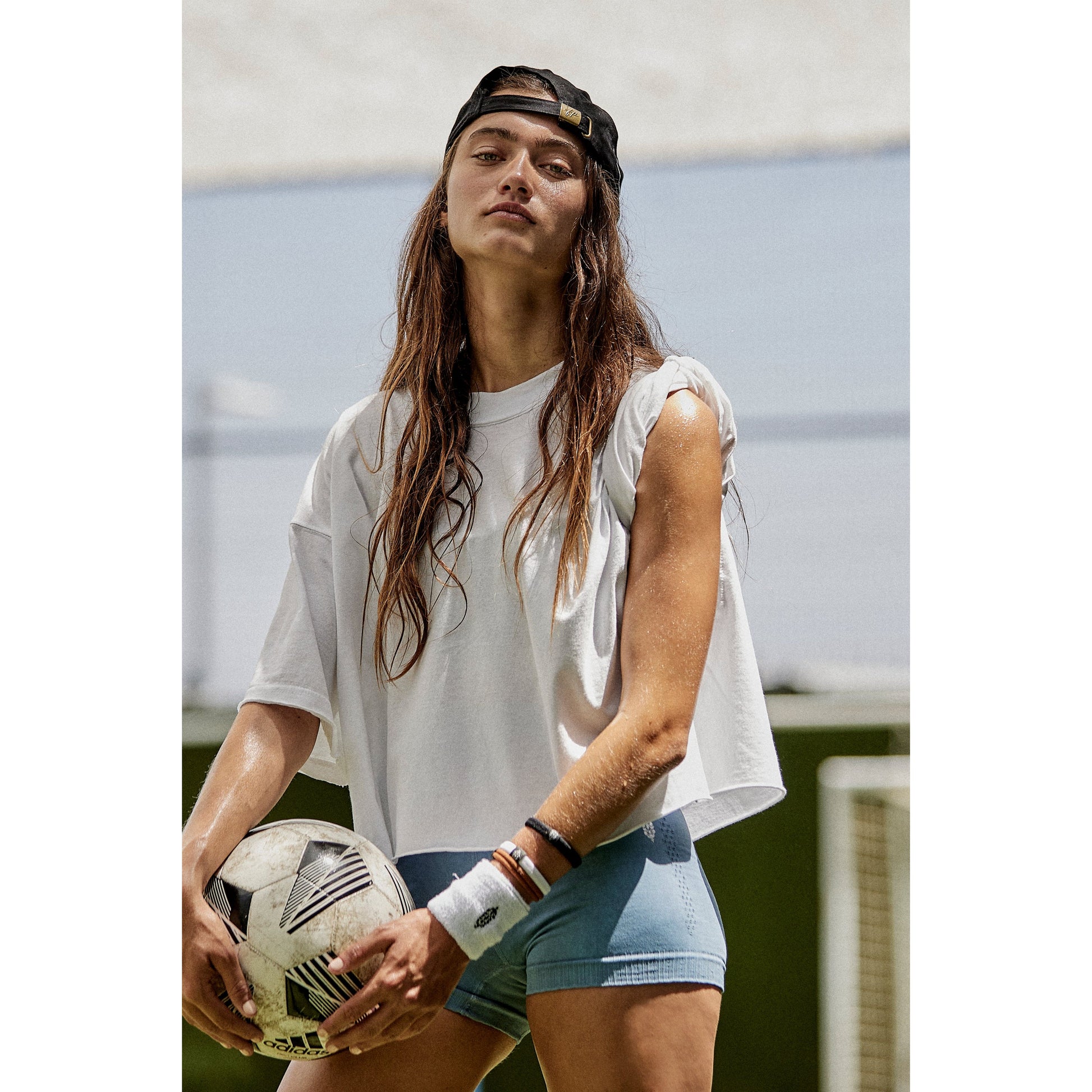 A woman in an oversized boxy fit Free People Movement Inspire Tee, White and denim shorts holding a soccer ball, wearing a black cap, standing on a field under a sunny sky.